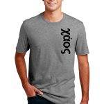 Young man in a gray T-shirt with the word "Xáos" vertically printed on it. He's standing casually with one hand in his pocket, wearing jeans, and smiling. The background is plain white.